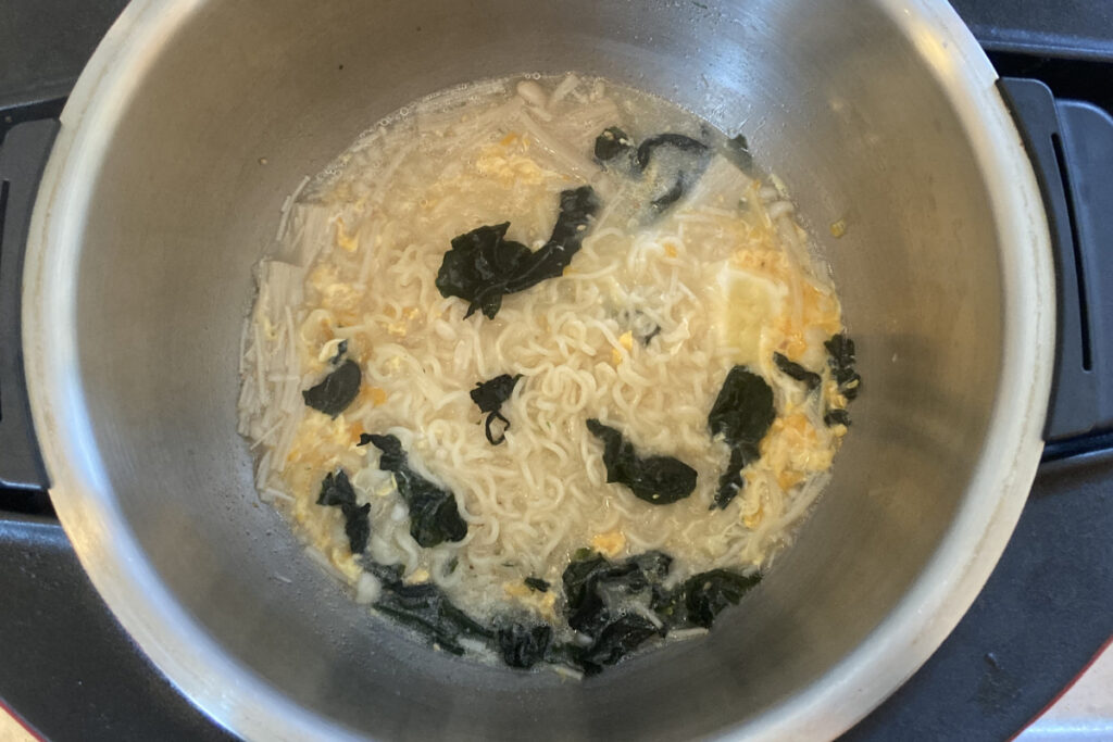 仕上げに溶き卵を回し入れたインスタントラーメンの様子。具材のワカメとえのき茸がスープに馴染み、卵がふわふわと固まり始めた、熱々の調理過程を伝えるクローズアップ写真。
