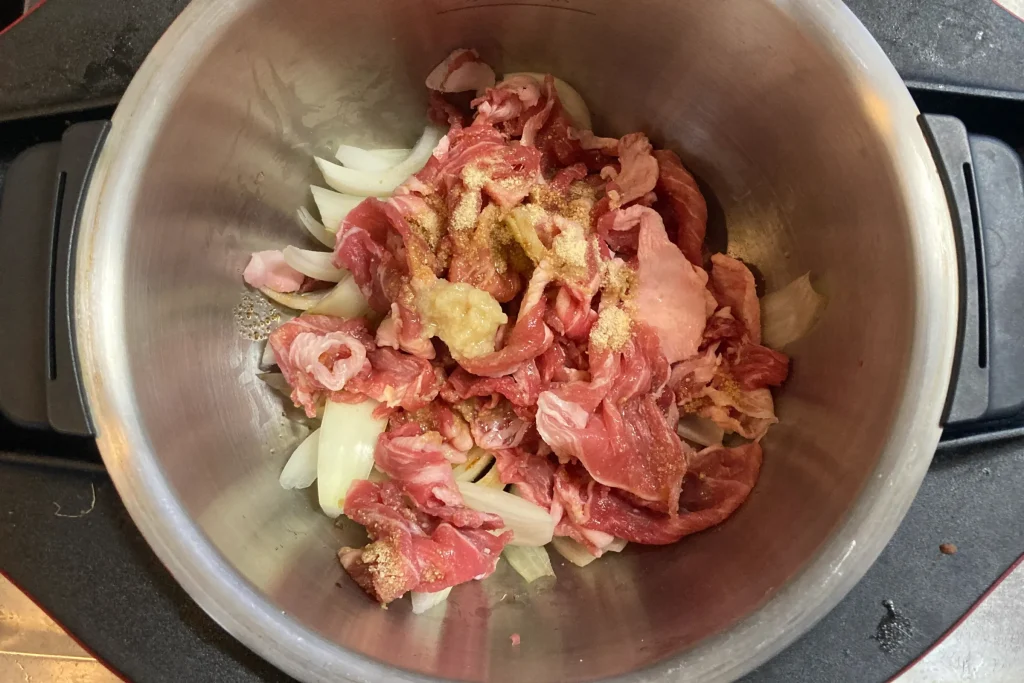 【基本・最適】 ステンレス製の鍋の中に、薄切りの生の豚肉とくし形切りにした玉ねぎが入り、その上にすりおろし生姜と茶色の粉末調味料が添えられている、調理前の様子。