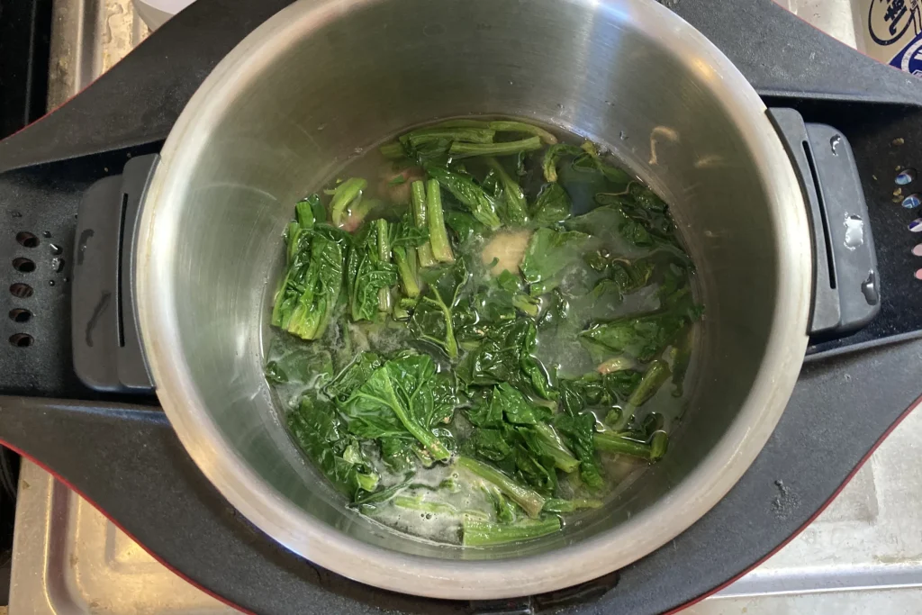 ステンレス製の両手鍋の中で、緑色の鮮やかなほうれん草が茹でられている様子。鍋底には少量の茹で汁が見え、ほうれん草の茎と葉が等間隔にカットされて浸かっている調理中の俯瞰写真