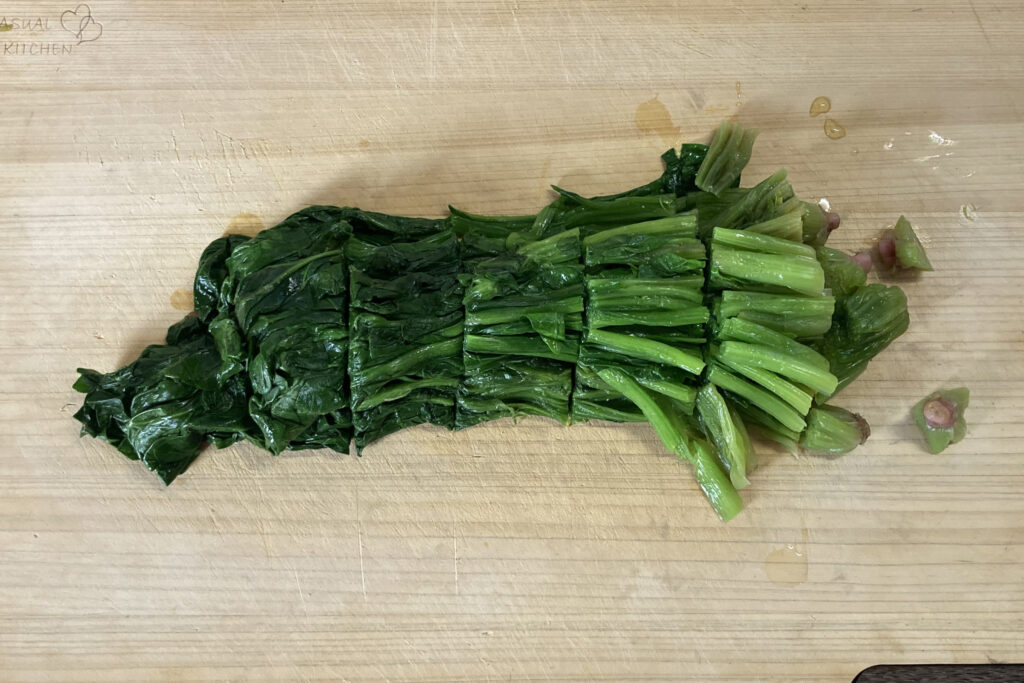 木製のまな板の上で、茹でた後のほうれん草が横一列に並べられ、食べやすい3〜4cm幅に等間隔で切り分けられている様子。右端には切り落とされたピンク色の根元の部分が置かれており、調理の下準備が整った状態。
