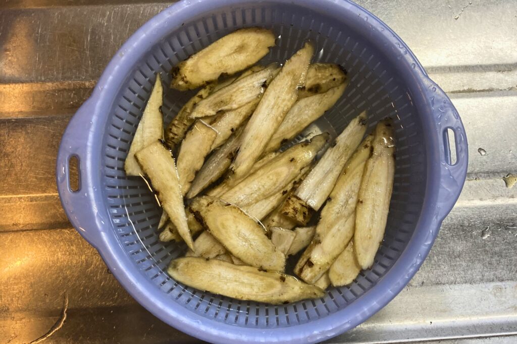 調理の下準備として、斜めに薄切りしたごぼうを青いザル付きのボウルに入れ、水にさらしてアク抜きをしている状態。