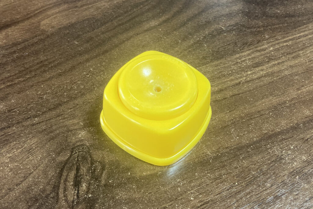 木目調のテーブルの上に置かれた、卵の殻に穴を開けるための黄色いプラスチック製キッチンツール。中央の円形のくぼみには小さな穴があり、卵を押し当てることで安全に針が飛び出し、ゆで卵を剥きやすくする仕組みを伝えています。