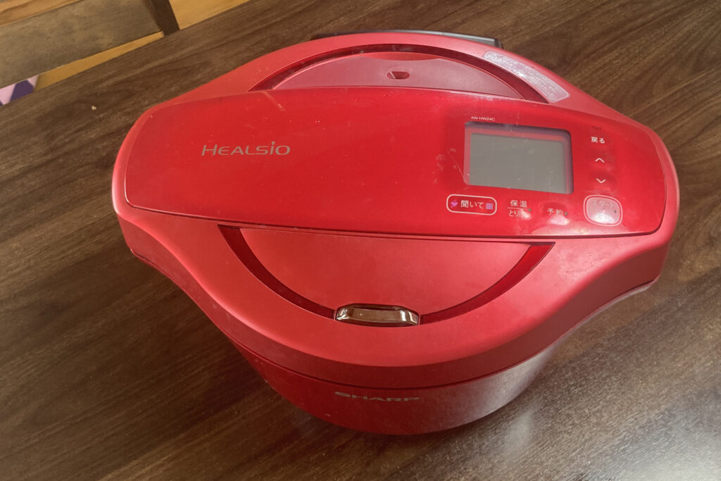 木目調のテーブルに置かれた、シャープ製の赤い水なし自動調理鍋「ヘルシオ ホットクック」。上部にはHEALSIOのロゴ、操作パネル、液晶画面があり、丸みを帯びた赤い筐体が特徴的なキッチン家電の外観。