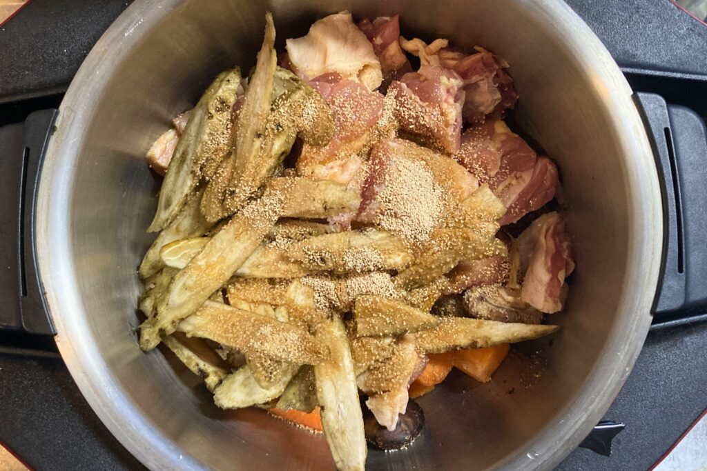 自動調理鍋のステンレス内釜に、カットされた鶏もも肉、ごぼう、にんじんなどの具材を入れ、上から出汁の粉末をかけた状態。