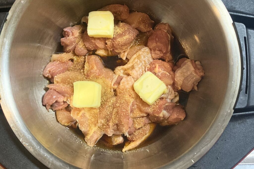 ホットクックの内鍋にカットした鶏もも肉と、にんにく、バター、醤油、酒、みりん、砂糖の調味料をすべて投入した状態。加熱前に全体をざっくりと混ぜ合わせ、味が均一に馴染むように準備を整えています。