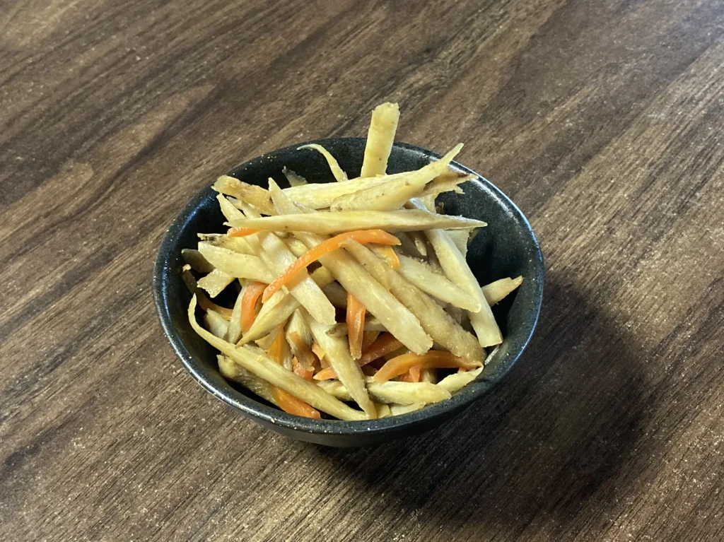 ホットクックで調理された、照り豊かなきんぴらごぼう。細切りにされたごぼうとにんじんが美しく混ざり合い、白いお皿に盛り付けられ、仕上げの白ごまがふりかけられた完成写真。