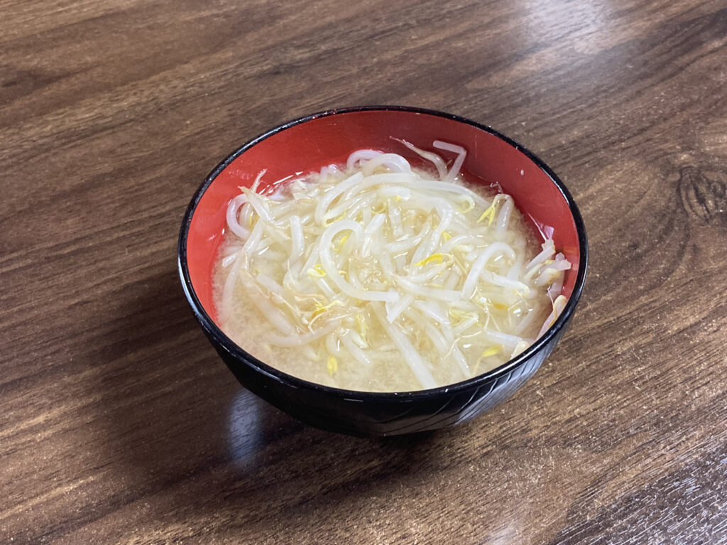 木目調のテーブルに置かれた、黒地に内側が赤のお椀に盛り付けられた「もやしの味噌汁」。ホットクックで調理され、もやしがたっぷり入った温かそうな仕上がり。