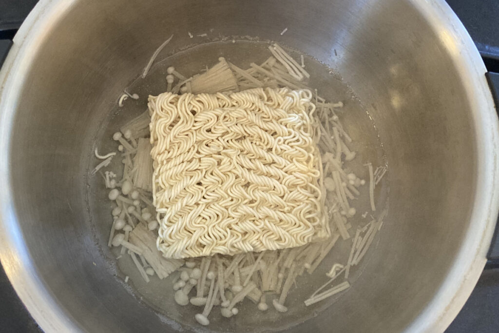 ホットクックで「うまかっちゃん」を作る工程。内鍋に水、えのき茸、インスタント麺の順で重ねて入れる。麺が水に半分ほど浸かっている、加熱調理を開始する直前の準備の様子。