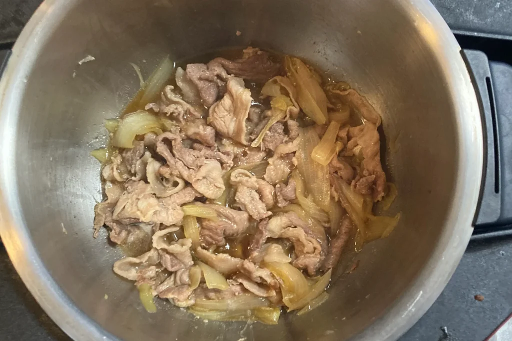 ステンレス製の内鍋の中で、薄切りの牛肉と飴色に透き通った玉ねぎが甘辛いタレで煮込まれている様子。煮汁が具材に染み込み、しっとりと仕上がった豚丼の具や肉豆腐のような煮物の調理風景です。