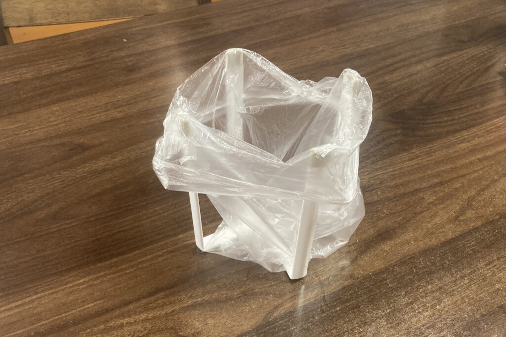 木目調のテーブルの上に置かれた、白いスチール製のポリ袋エコホルダー。透明なビニール袋が四隅の支柱に掛けられており、調理中のゴミ入れとして自立している様子を捉えた斜め上からの写真。