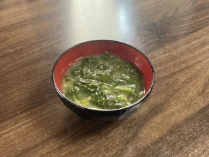 木目調のテーブルに置かれた、黒地に内側が朱色の汁椀に盛り付けられたほうれん草の味噌汁。たっぷりの青菜が汁に浸かり、家庭的な温かさが伝わる料理写真。