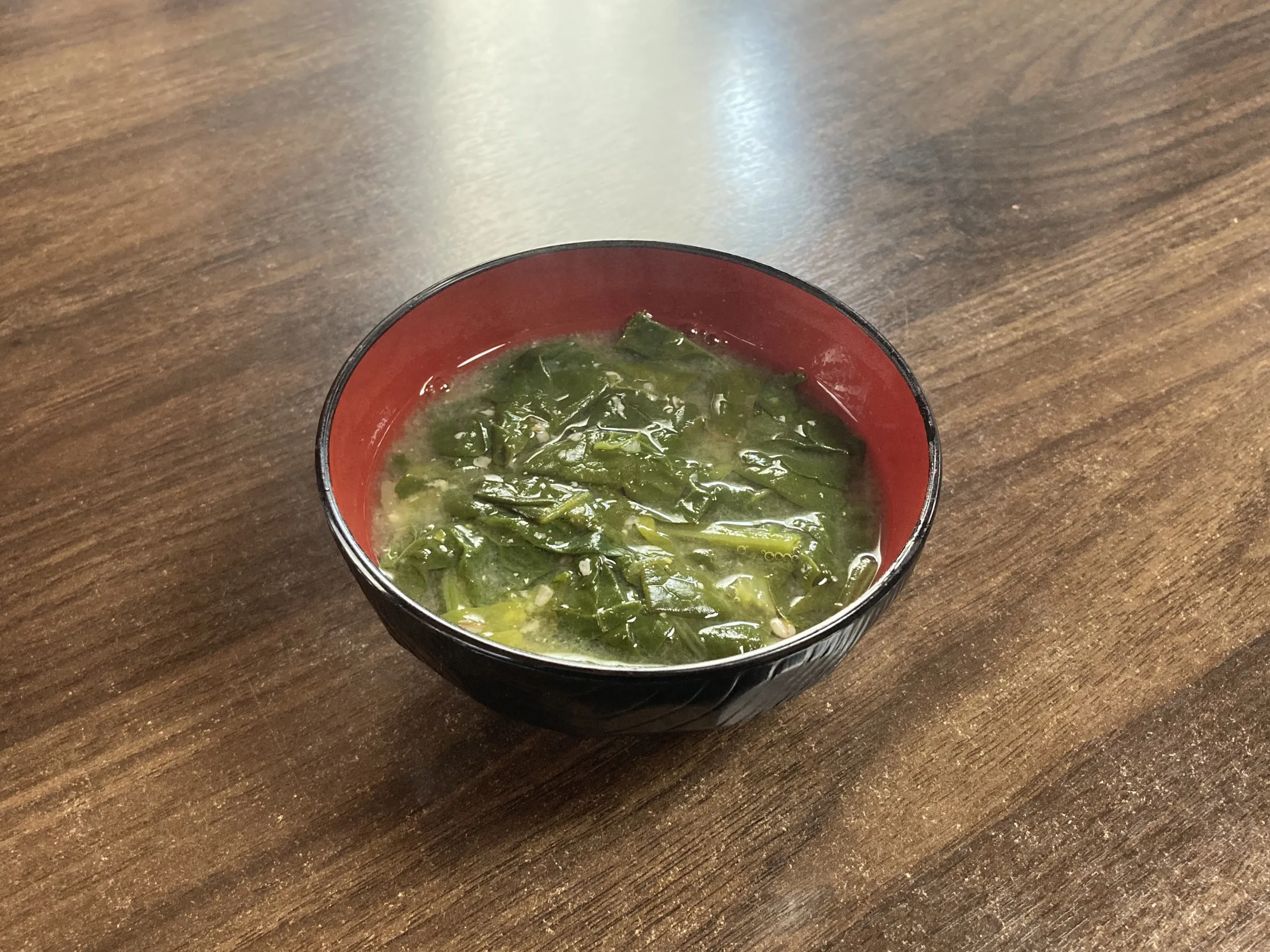 木目調のテーブルに置かれた、黒地に内側が朱色の汁椀に盛り付けられたほうれん草の味噌汁。たっぷりの青菜が汁に浸かり、家庭的な温かさが伝わる料理写真。