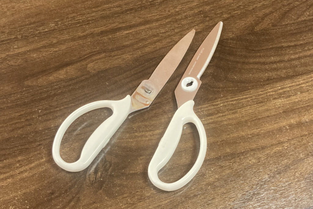 木目調のテーブルの上に置かれた、白いハンドルのプラス製キッチンバサミ。洗浄や手入れのために左右の刃がジョイント部分から取り外され、2つのパーツに分かれた状態を斜め上のアングルから撮影している。