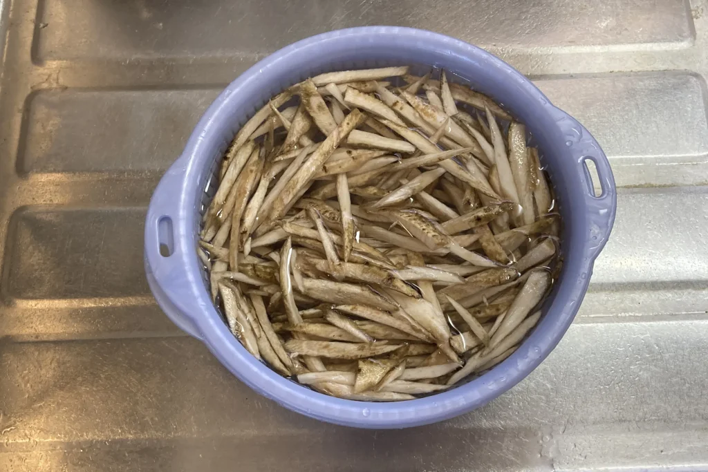 水を入れた薄紫色のプラスチックボウルの中で、ささがきにしたごぼうをアク抜きのために浸している、キッチンのシンクでの調理風景。
