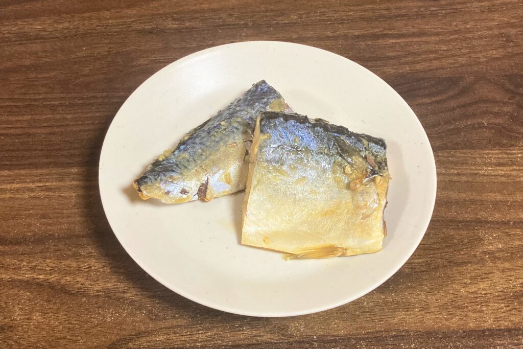 木目のテーブルの上に置かれた白い丸皿に、サバの味噌煮が2切れ盛り付けられている。皮目が上を向いている。
