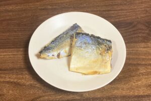 木目のテーブルの上に置かれた白い丸皿に、サバの味噌煮が2切れ盛り付けられている。皮目が上を向いている。