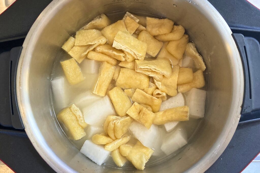 ステンレス製の鍋の中で、いちょう切りにした大根と、食べやすい大きさに切った油揚げが、ひたひたのだし汁で煮られている様子。