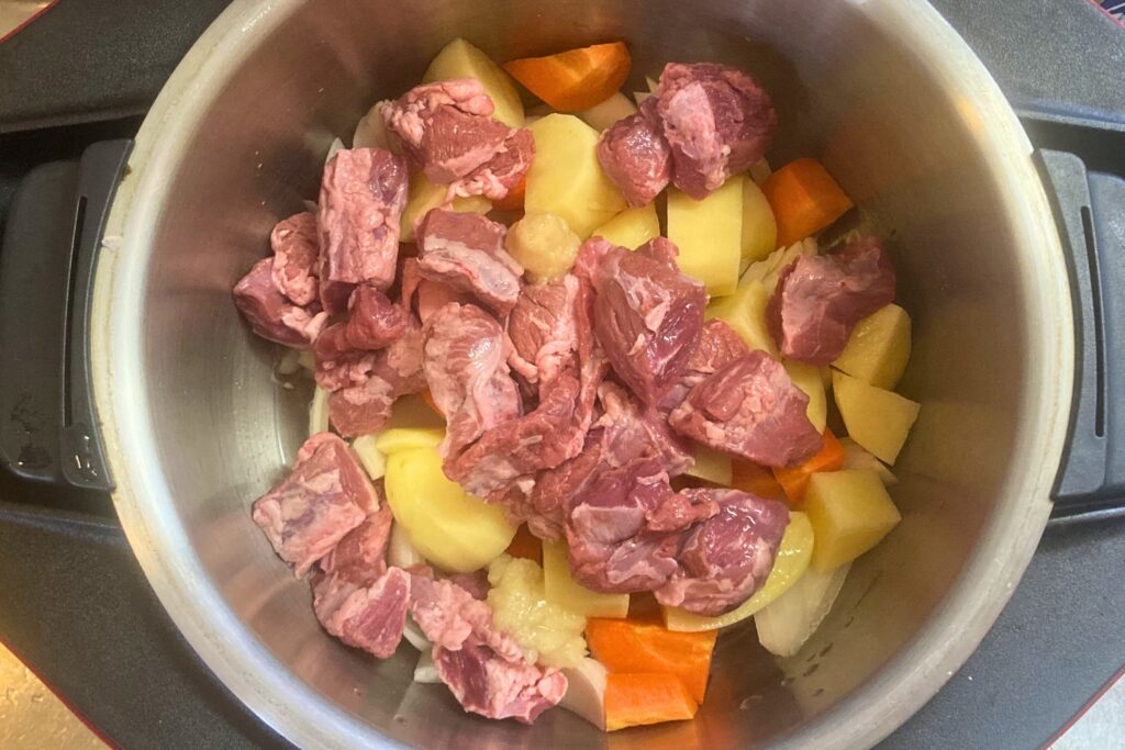自動調理鍋のステンレス製の内鍋に、調理前の生の豚肉、大きくカットされたじゃがいも、にんじん、玉ねぎが山盛りに詰め込まれている様子です。これから加熱調理が始まります。