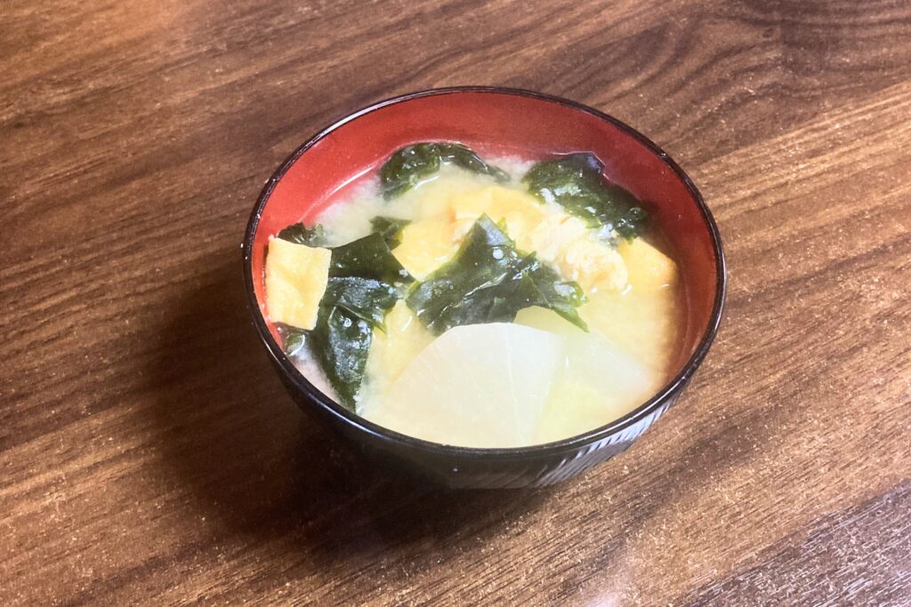 木目調のテーブルの上に置かれた黒と赤の漆器風のお椀に、大根、油揚げ、わかめがたっぷり入った具だくさんの温かいお味噌汁。