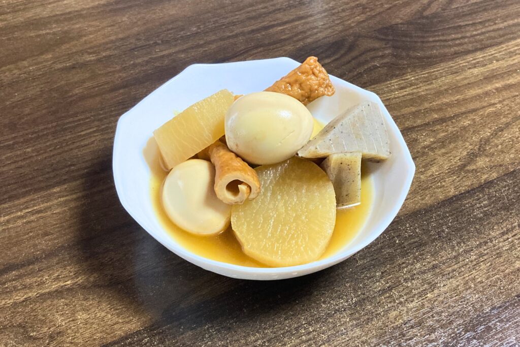 白い六角形の器に盛り付けられた完成したおでん。出汁の色が染みた大根、ゆで卵、こんにゃく、ごぼう巻、ちくわが彩りよく並ぶ。