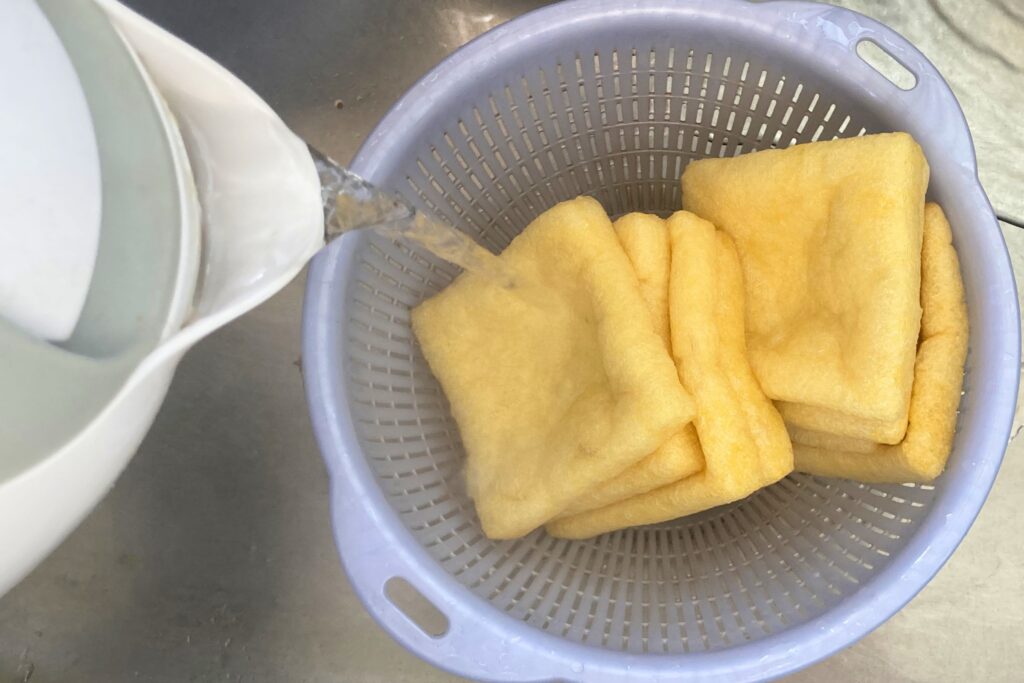 水色のプラスチック製ザルに入れられた数枚の油揚げの上から、白い電気ケトルを使って熱湯を注ぎ、油抜きをしている調理工程。