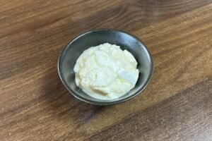 ダークブラウンの木目調テーブルの上に置かれた、質感のある黒い小鉢に入った、濃厚でクリーミーなギリシャヨーグルト。表面にはスプーンですくったような凹凸が見られる。