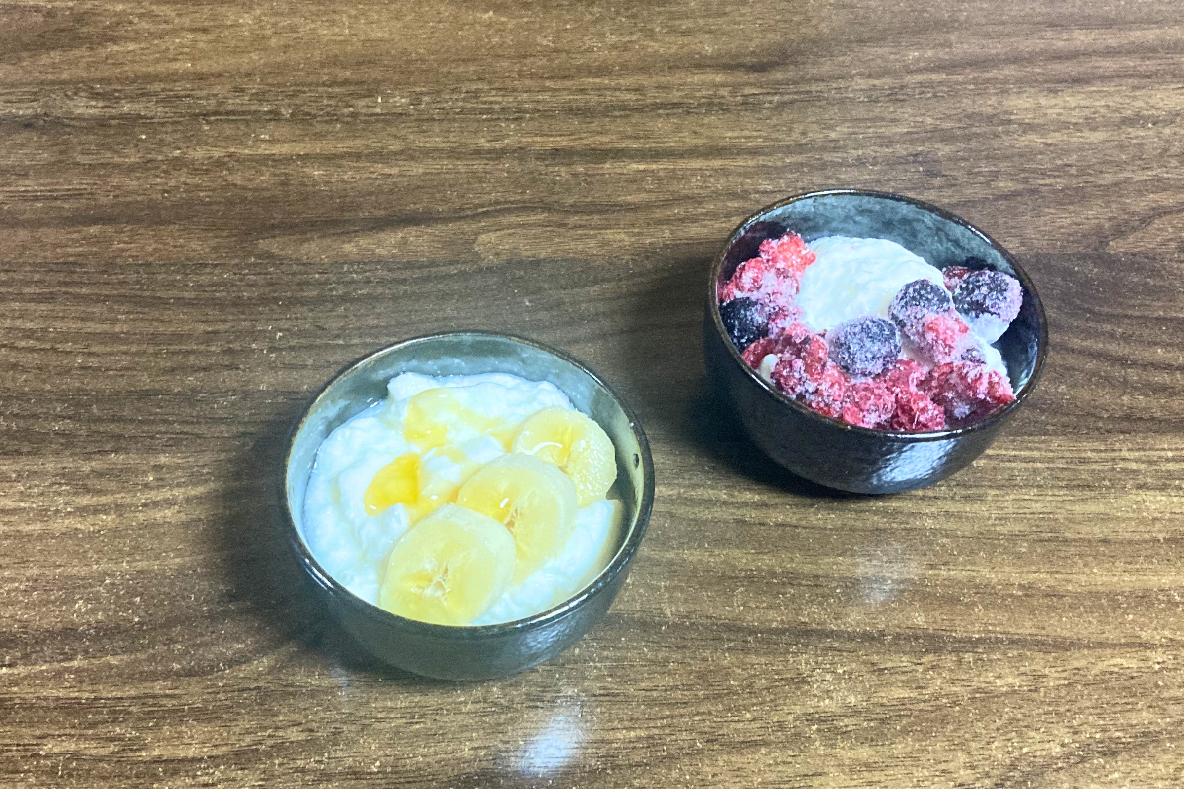 木目調のテーブルに置かれた2つの小鉢。左はバナナと蜂蜜、右はフローズンベリーがトッピングされた自家製ヨーグルト。