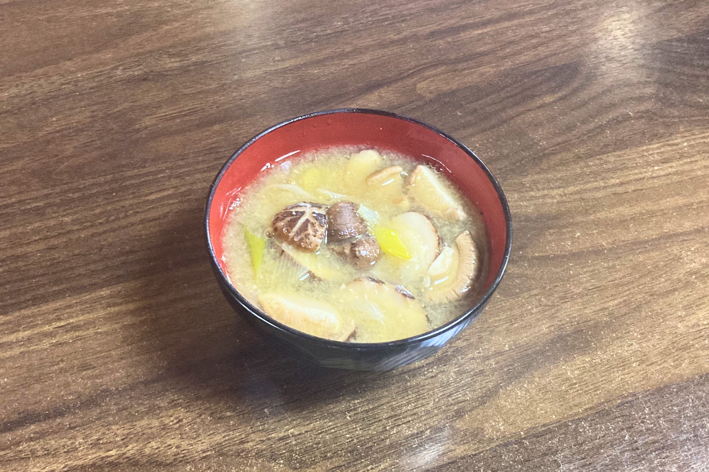 赤茶色の漆器風のお椀に盛り付けられた、しいたけと長ねぎの温かい味噌汁。木目のテーブルの上に置かれた完成写真。