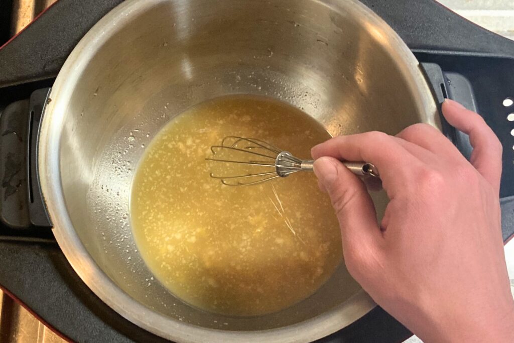 電気圧力鍋の内鍋で、黄色い煮汁を小さな泡立て器を使ってかき混ぜている様子。右手が写っている。