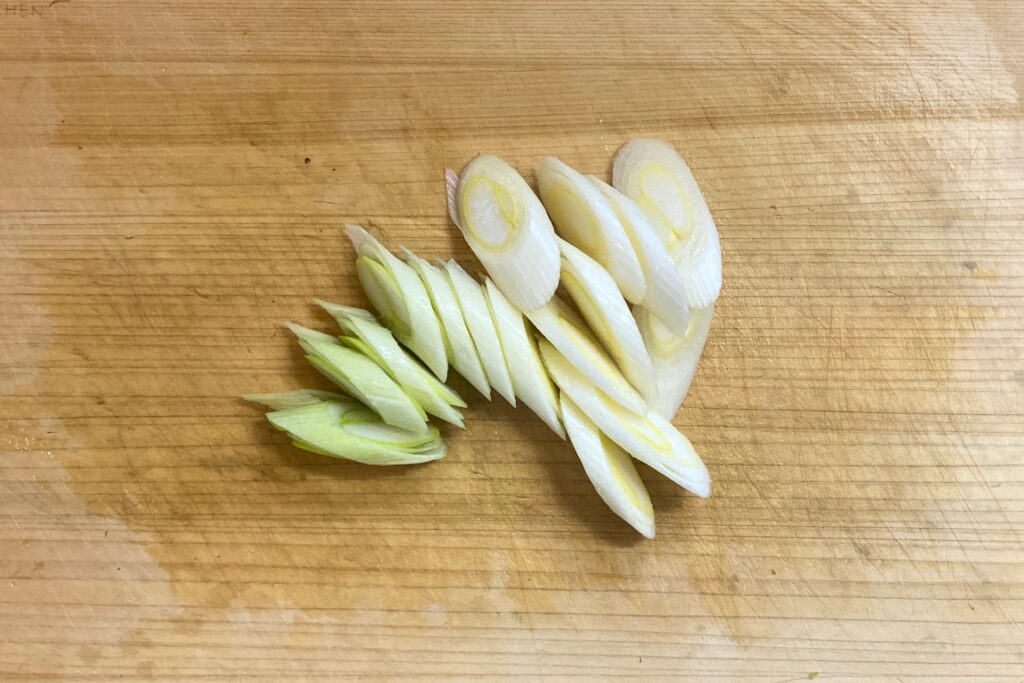 木目のまな板の上で、約1センチ幅の斜め切りにされた白ねぎ（長ねぎ）。白い部分から青い部分へのグラデーションが見える。