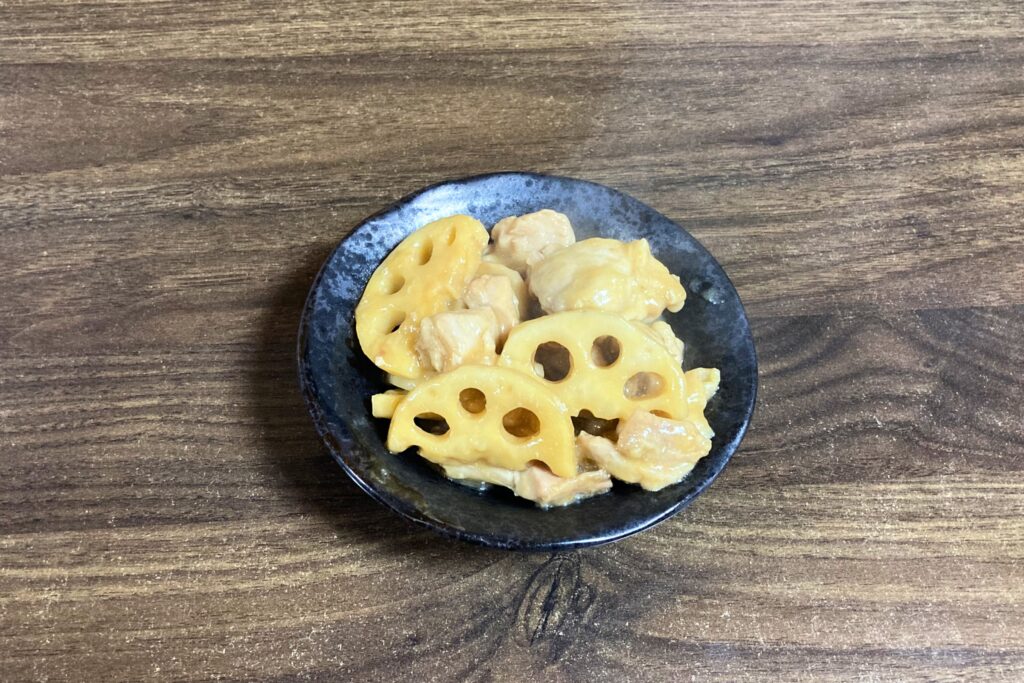 木目調のテーブルの上で、黒い和風の小鉢にこんもりと盛り付けられた、完成した鶏肉とレンコンの照り焼き風炒め物。