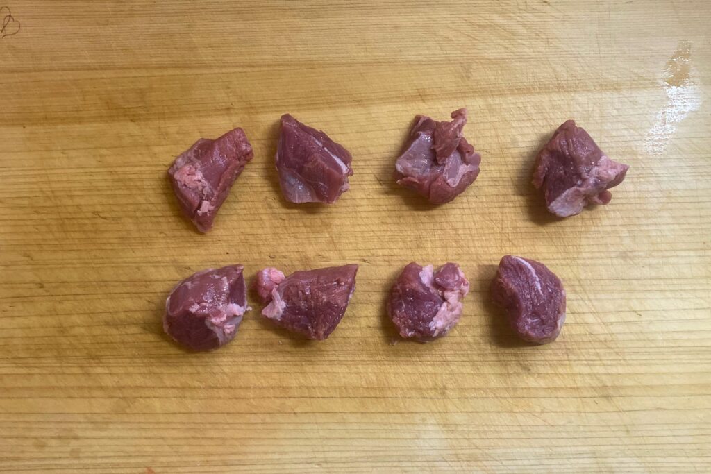 木製のまな板の上に、カレーなどの調理用として一口大に切り分けられた生の豚肉が8切れ、等間隔に並べられています。赤身と脂身が混ざった新鮮な状態がはっきりと確認できます。