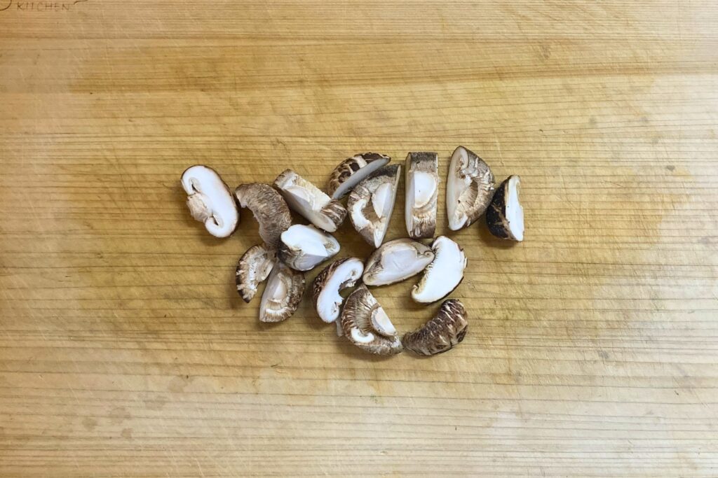 木目のまな板の上で、石づきを切り落としてから食べやすい大きさのそぎ切りにされた、新鮮な生しいたけのカット写真。