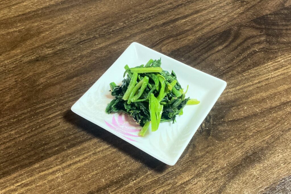 小さな白皿に盛られた小松菜のお浸し。醤油とごまで和えられた緑の野菜が、木目調のテーブルの上にあります。皿の隅にピンクの模様。