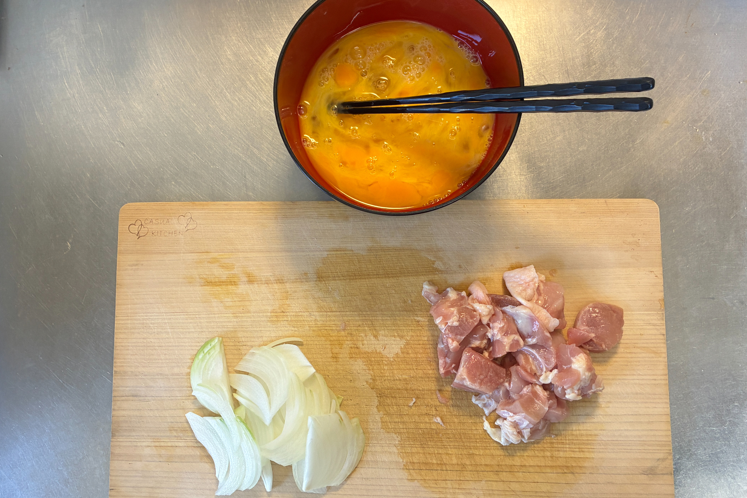 木製のまな板の上に一口大に切った鶏もも肉と薄切りにした玉ねぎが並び、上部には黒い箸を添えた赤い器に溶きほぐした生卵が用意されている調理前の下準備の様子。