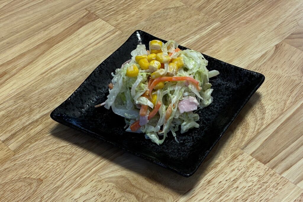 黒い角皿に盛り付けたコールスロー。マヨネーズで和えたキャベツとにんじんにハムが混ざり、上にコーンがのっている