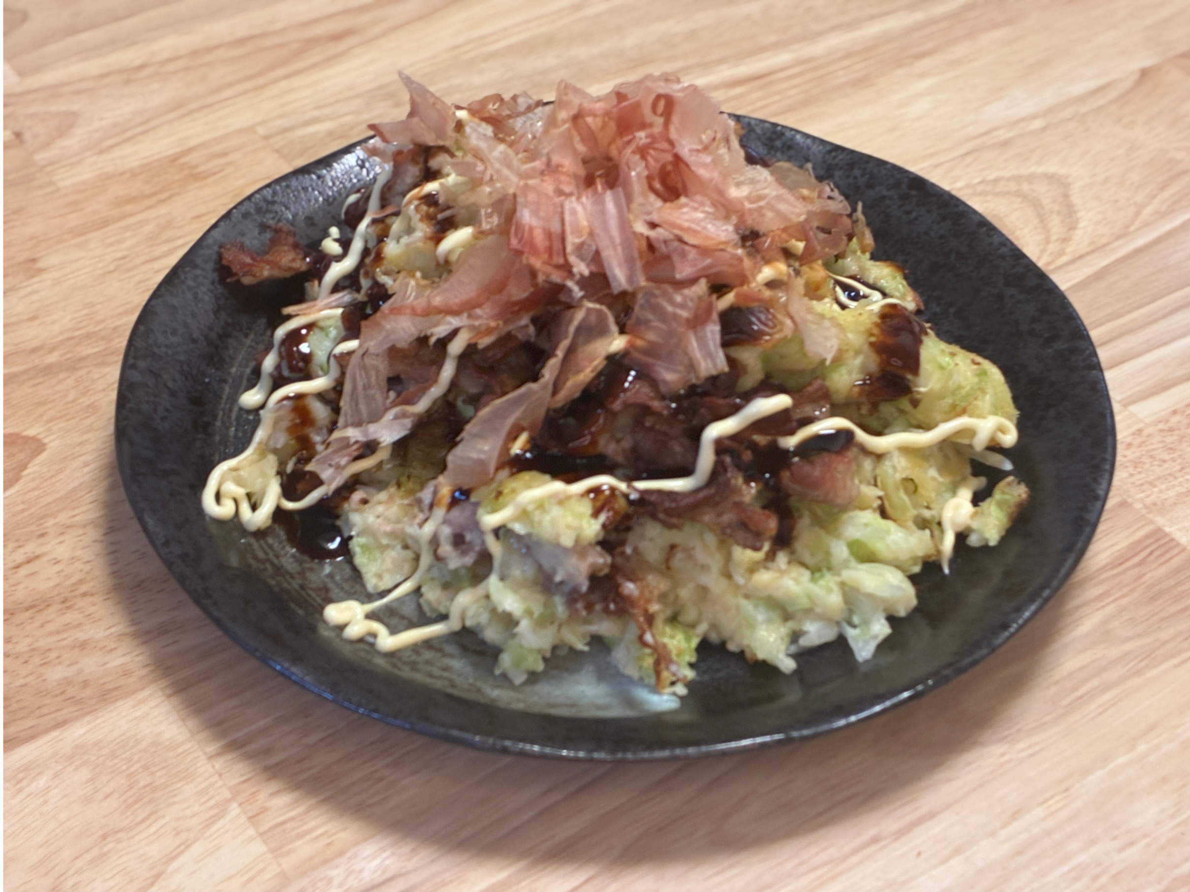 ヘルシオで焼いたお好み焼き。黒い皿に盛り、ソースとマヨネーズ、かつお節をのせている