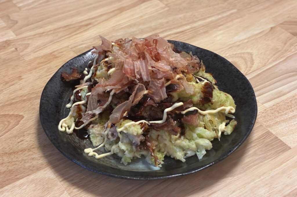 黒い皿に盛ったお好み焼き。ソースとマヨネーズをかけ、かつお節をたっぷりのせている