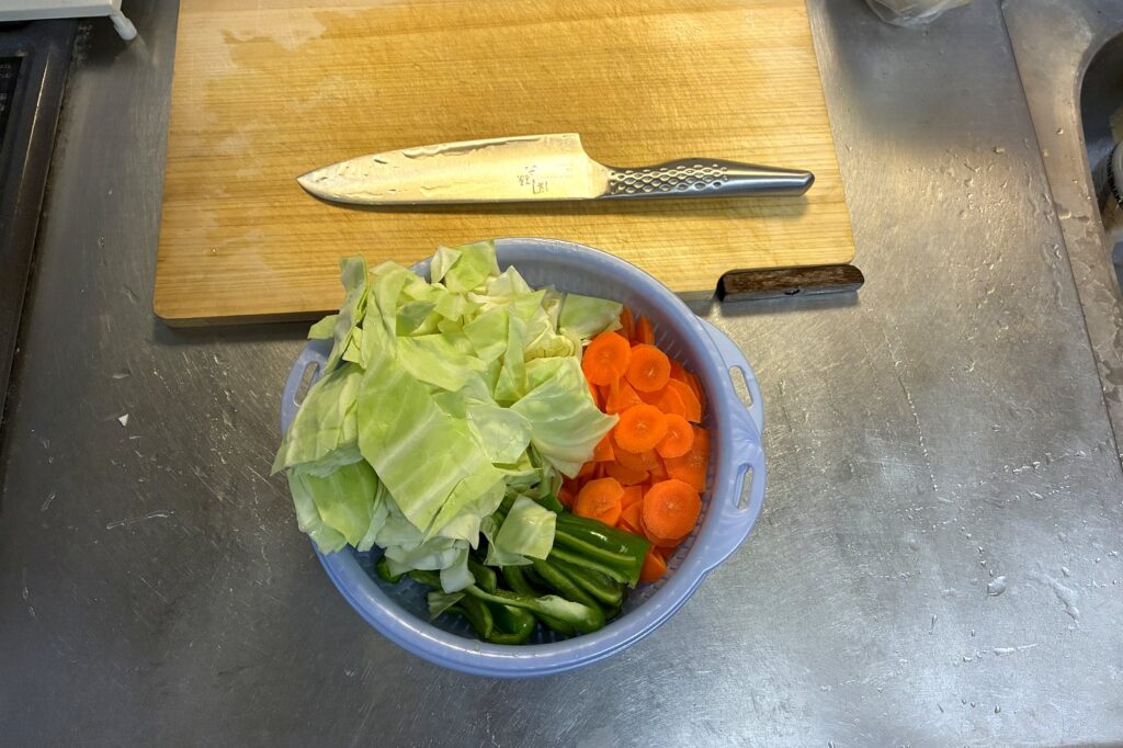 ざく切りにしたキャベツと輪切りのにんじんなどの野菜をボウルにまとめた下ごしらえの状態。奥にまな板と包丁が置かれている。