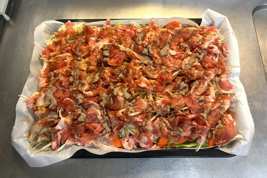 野菜の上に豚こま肉を重ならないように広げ、その上から付属の粉末ソースを全体に振りかけた状態。