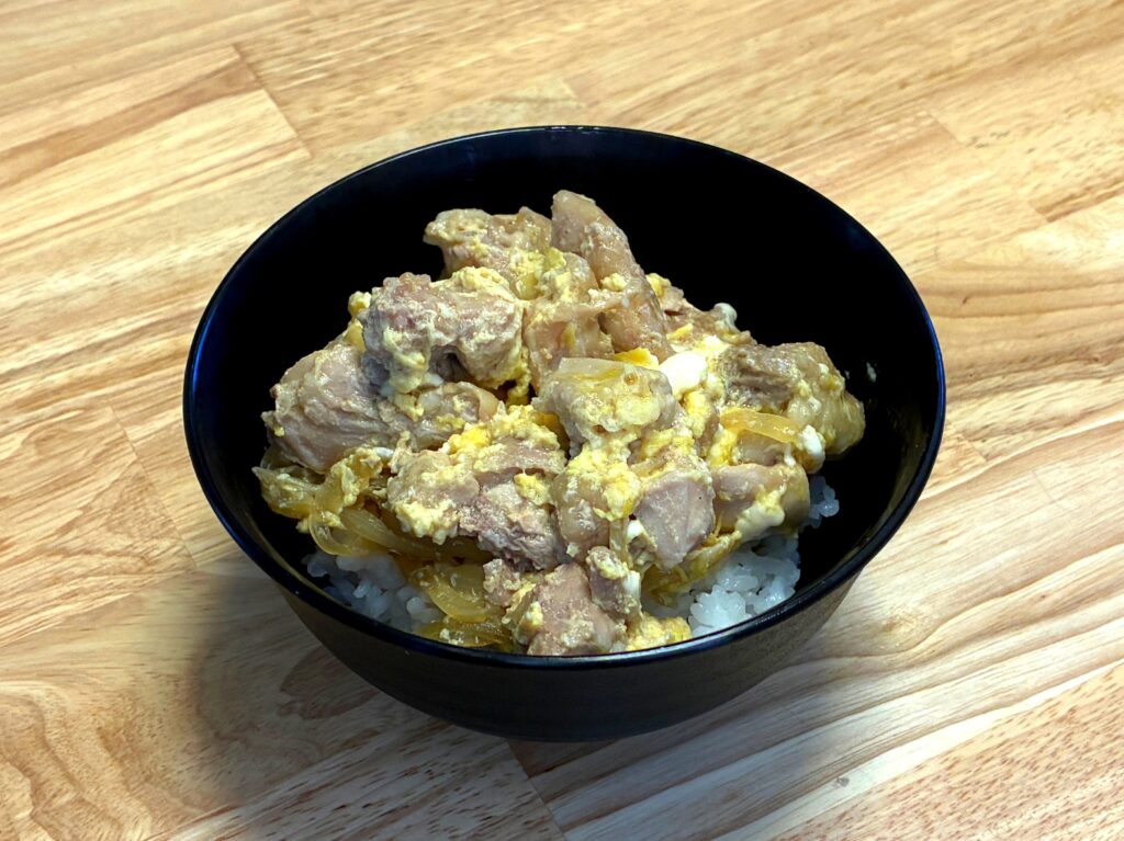 木目調のテーブルの上に置かれた黒いどんぶりに、ふんわりと卵でとじられた鶏もも肉と玉ねぎがご飯の上にたっぷりと盛り付けられたホットクックで作る親子丼の完成写真。