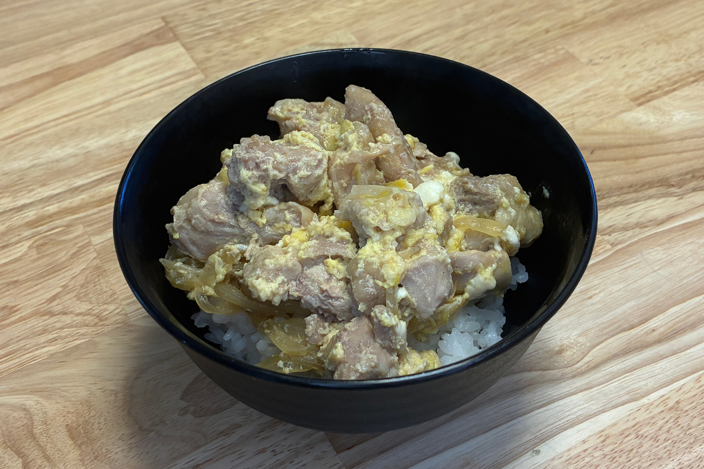木目テーブルの上の黒いどんぶり鉢に、炊き立てのご飯をよそい、ホットクックで煮上げたふんわり卵と大ぶりの鶏もも肉、玉ねぎを汁ごとふんだんに盛り付けた完成写真。