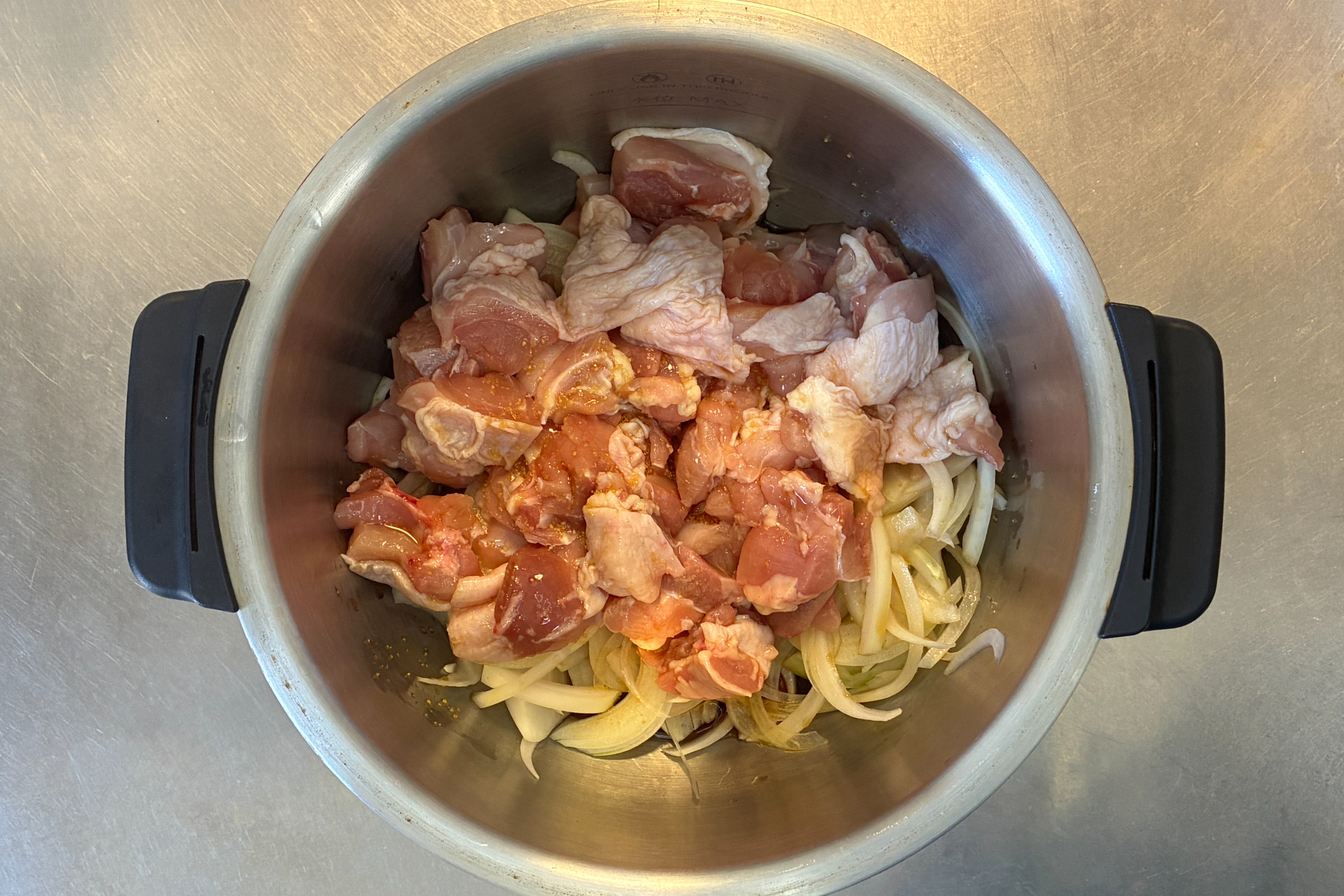 ホットクックのステンレス内鍋の底に薄切り玉ねぎを敷き詰め、その上に生の鶏もも肉を重ならないように広げてのせ、調味料を回しかけた調理開始直前の状態。