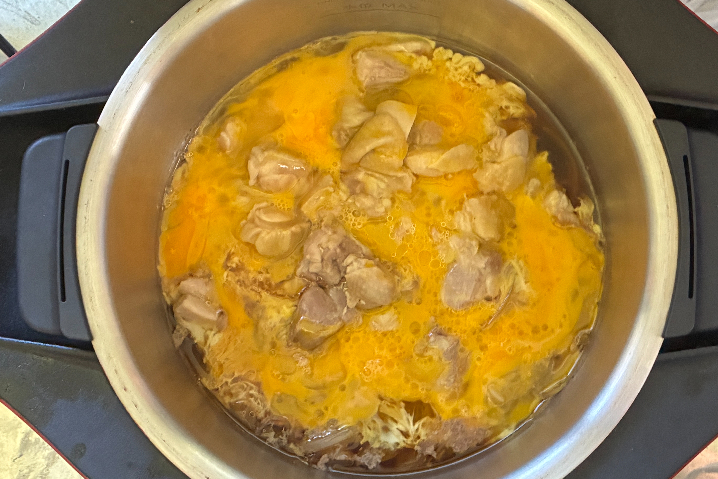 加熱が終わった直後のホットクック内鍋へ、黄色い溶き卵を全体に回し入れた様子。熱々の鶏肉と玉ねぎの上に生の卵液がふんわりと広がっている調理工程。
