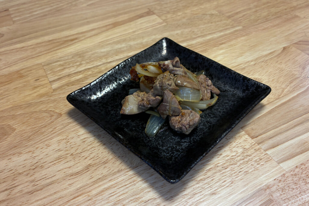 黒い正方形の和皿に、甘辛だれを絡めて照りよく盛り付けられた鶏レバーとハツの甘辛しょうが蒸しの完成品。しっとり柔らかなレバーと甘い玉ねぎが食欲をそそる仕上がり。